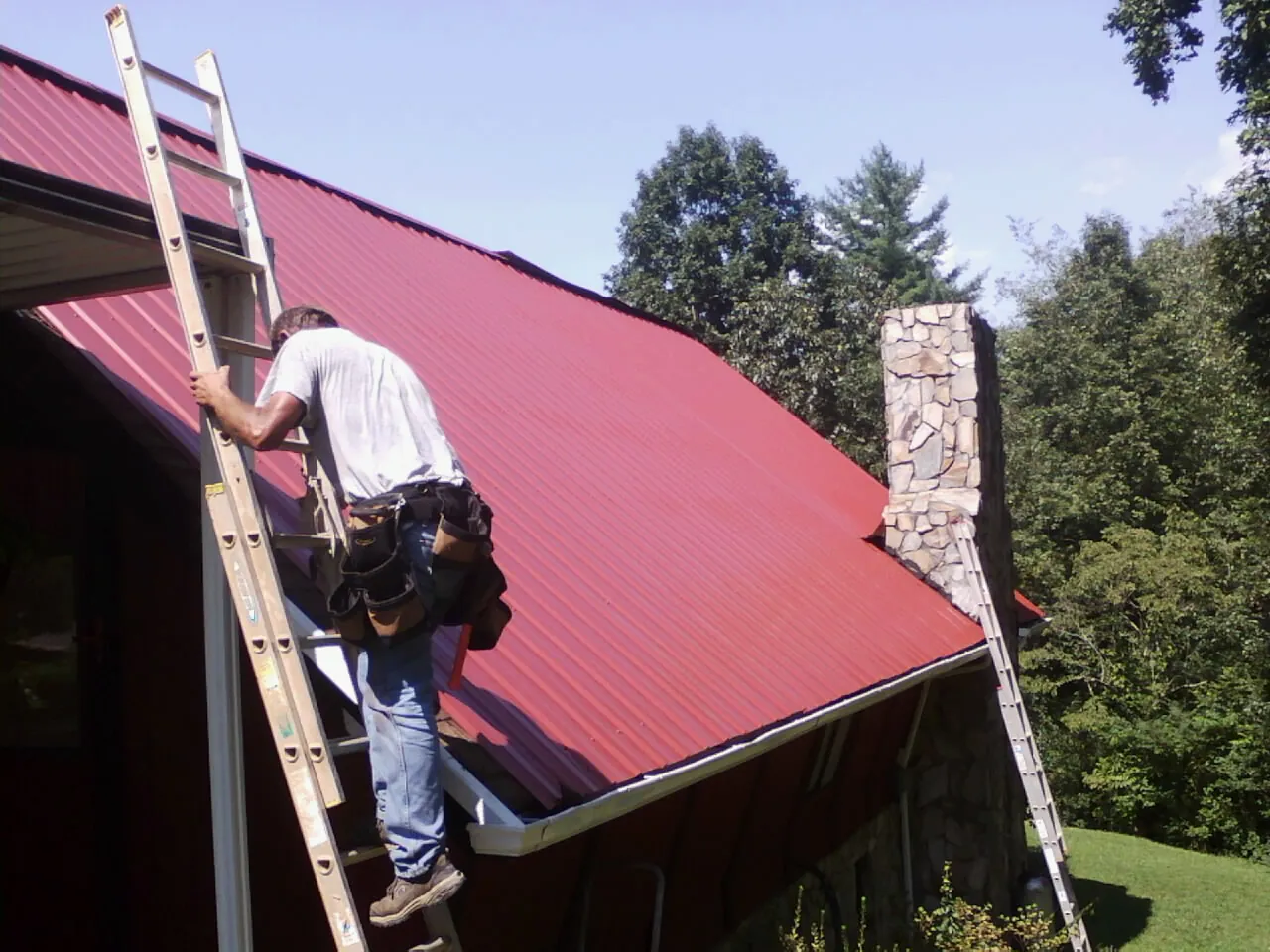 Roofer on a ladder beside a red metal roof and stone chimney — Soffit & Fascia Repair by Roofing Brothers in Bentley